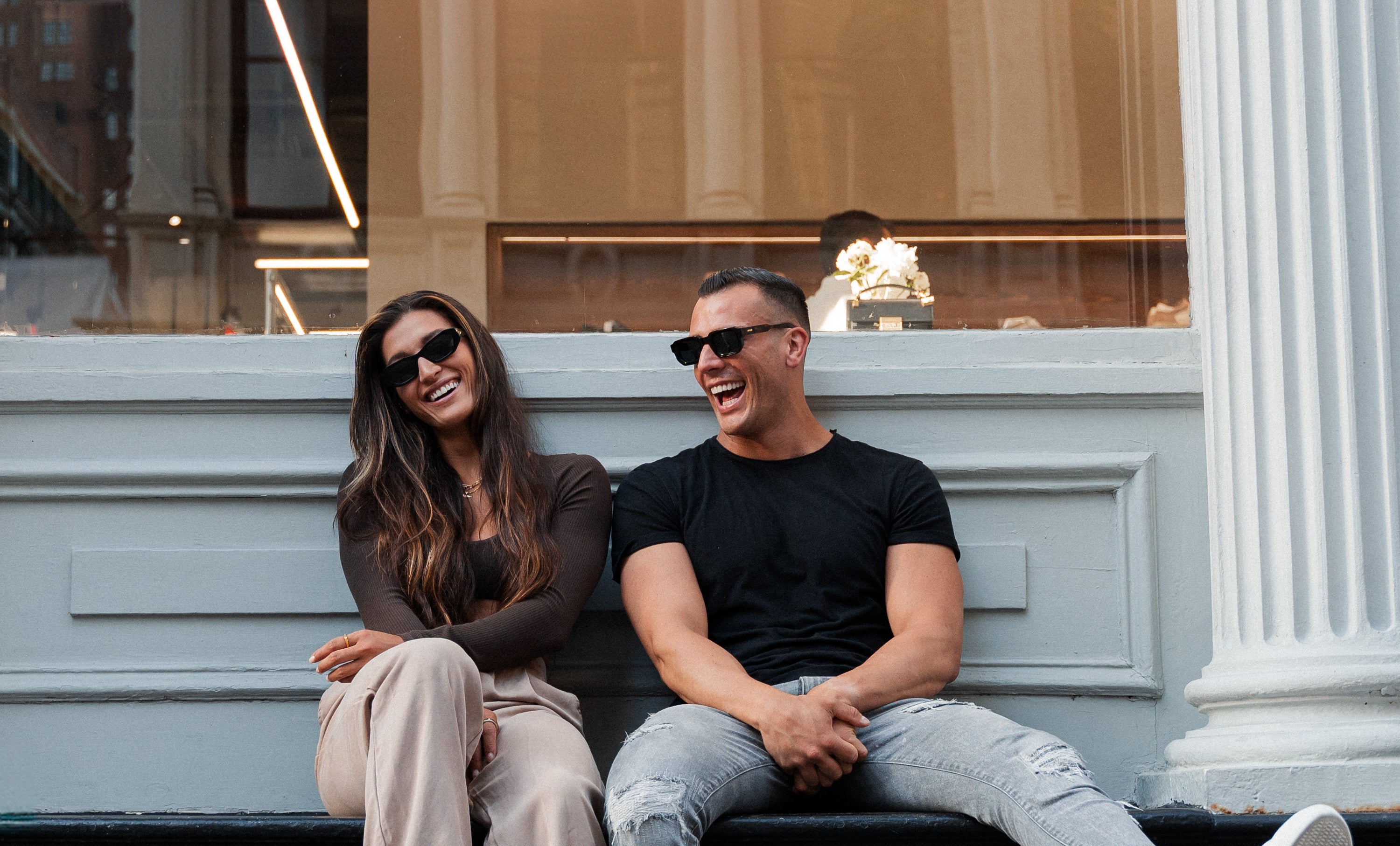 A couple wearing the A'PAIR pyrus and bartletts all-nighter sunglasses in black and laughing in the streets of New York